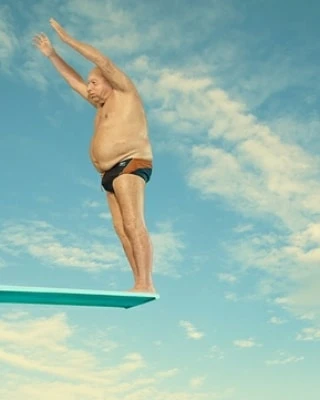 Man prepares to dive from a diving board against a blue sky with clouds. He is wearing a swimsuit and is poised with arms raised.